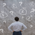 Pessoa olhando para um quadro com pontos de interrogação e cifrões, representando as dúvidas de investimentos para iniciantes.